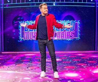 Host on a colorful game show stage, arms open, bright lights and "Musical Chairs" sign behind.