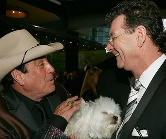 Two men in suits smiling at each other; one holds a small white dog.