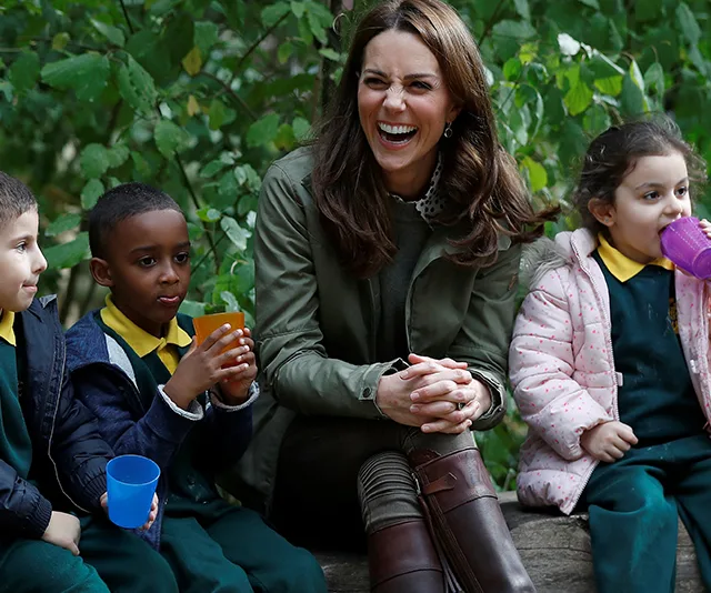 Kate Middleton laughing with three children, holding colorful cups, sitting outdoors in a green area.