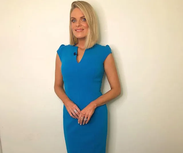 Woman in a blue dress standing against a plain backdrop, smiling with hands clasped in front.
