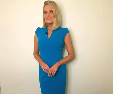 Woman in a blue dress standing against a plain backdrop, smiling with hands clasped in front.