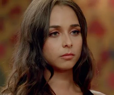 A woman with long dark hair looks serious against a blurred warm-toned background.