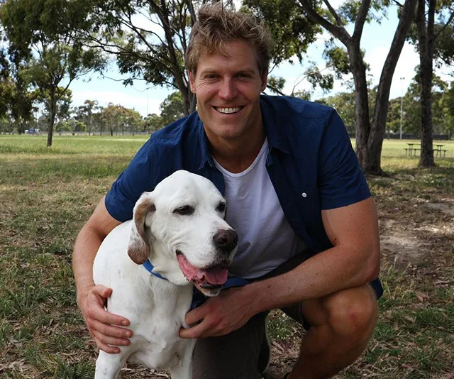 Man kneeling and hugging a white dog in a sunny park, both looking at the camera, with trees in the background.