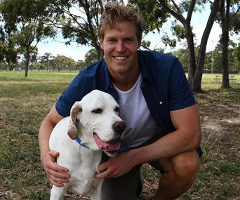 Man kneeling and hugging a white dog in a sunny park, both looking at the camera, with trees in the background.
