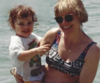 Woman in sunglasses holds a smiling child in water, both casually dressed.