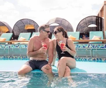 Couple sitting by the pool, smiling and holding red drinks, with lounge chairs and pods in the background.