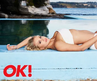 Woman in white swimsuit lounging by a pool, with coastal scenery in the background.