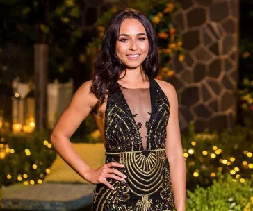 Woman in an elegant black and gold dress, smiling, standing outdoors with decorative lights in the background.