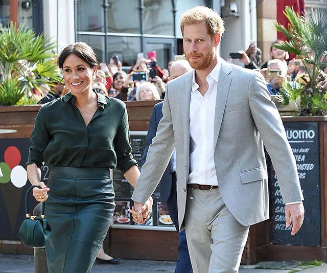 Couple walking hand-in-hand, surrounded by onlookers with cameras, in an outdoor setting.
