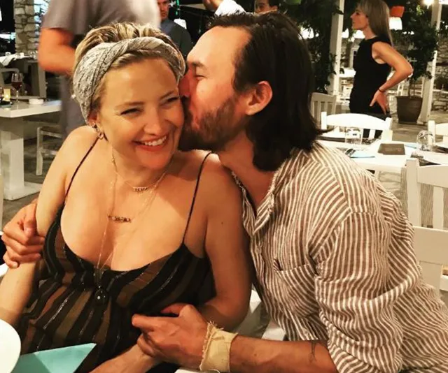 A couple embraces at a restaurant; the man kisses the woman's cheek, both smiling warmly.