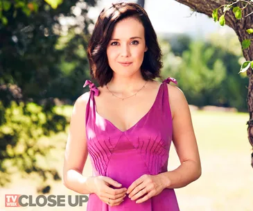 A woman in a magenta dress stands outdoors, surrounded by greenery, with a calm expression.