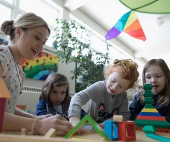 Teacher and three children interacting at a table with colorful blocks and decorations in a bright classroom.