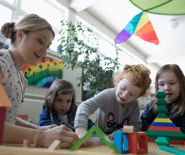 Teacher and three children interacting at a table with colorful blocks and decorations in a bright classroom.