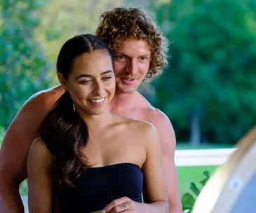 A couple stands closely together, smiling, with a blurred green background.