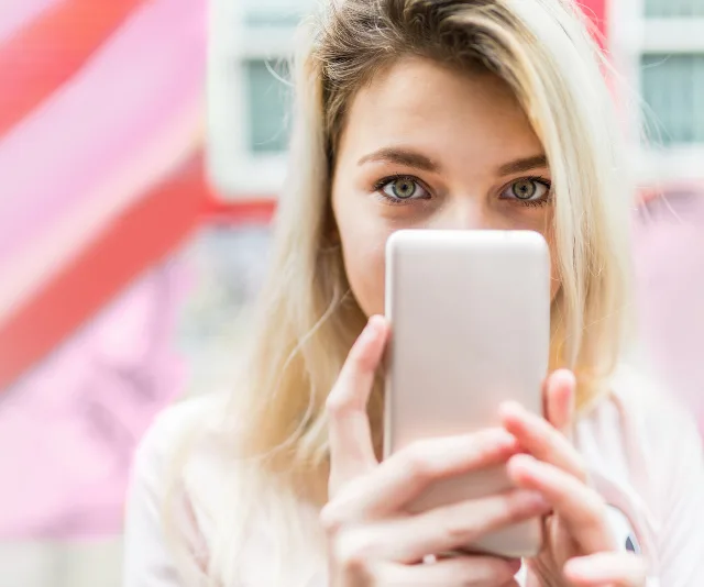 A woman with blonde hair holds a smartphone, capturing a selfie against a vibrant, blurred background.
