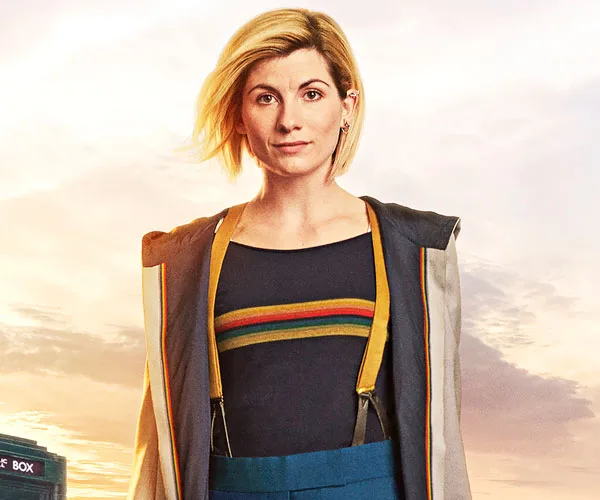 Jodie Whittaker as the Doctor in "Doctor Who" stands confidently, wearing her iconic rainbow-striped shirt and coat.
