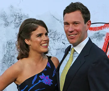 A woman in a floral dress smiling at a man in a suit with a patterned tie, posing in front of an abstract backdrop.