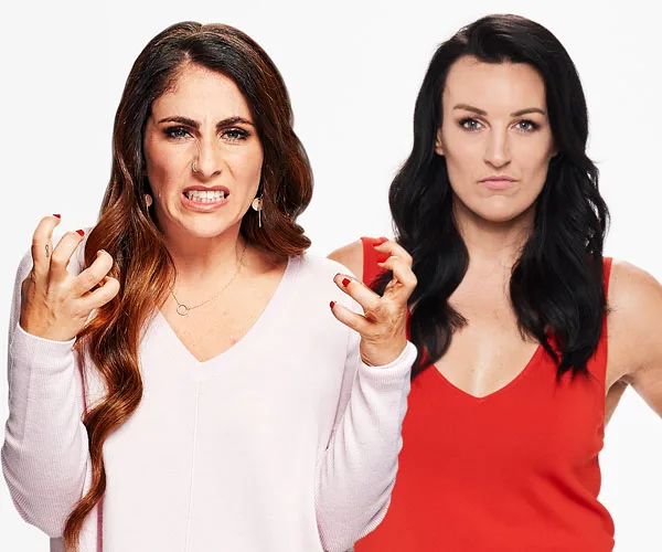 Two women standing, one expressing anger, the other neutral, in studio background.