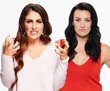 Two women standing, one expressing anger, the other neutral, in studio background.
