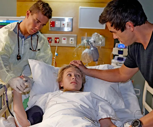 A woman lies in a hospital bed with a doctor holding her hand. Another person stands next to the bed, touching her forehead.