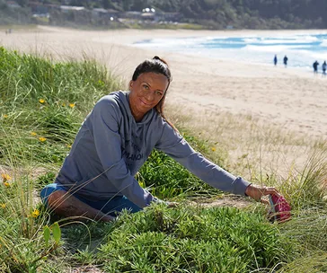 Person in hoodie planting on a beach dune with ocean in background.