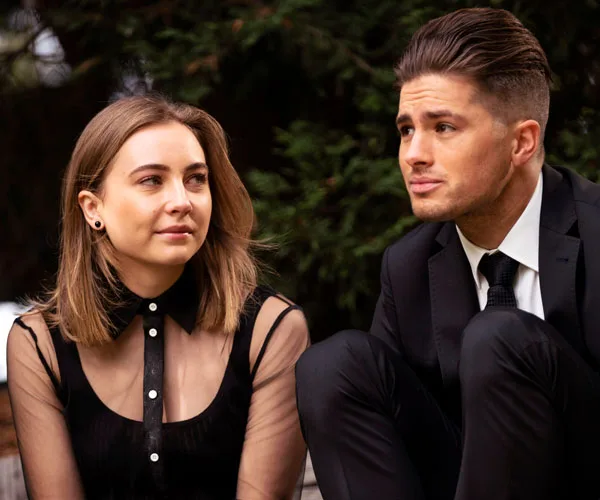 Two individuals sitting outdoors; a woman in a black dress and a man in a suit, both looking at each other.