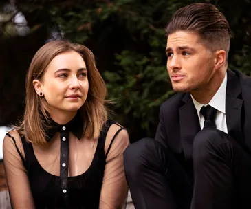 Two individuals sitting outdoors; a woman in a black dress and a man in a suit, both looking at each other.