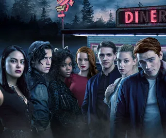 Cast of Riverdale standing in front of a diner at night, with neon signs and trees in the background.