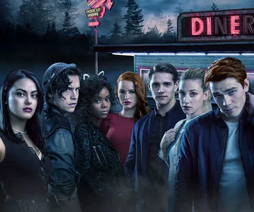 Cast of Riverdale standing in front of a diner at night, with neon signs and trees in the background.