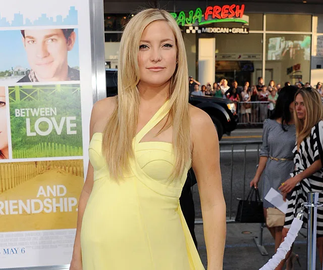 A woman in a yellow dress at a film premiere, standing next to a movie poster that reads "Between Love and Friendship."