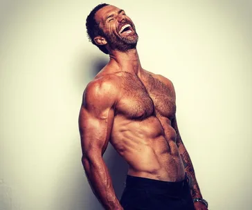 Man with a muscular build and beard laughing, shirtless against a plain backdrop.