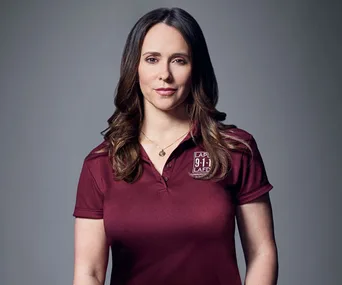 Jennifer Love Hewitt in maroon 9-1-1 uniform, posing confidently with soft smile, dark grey background.