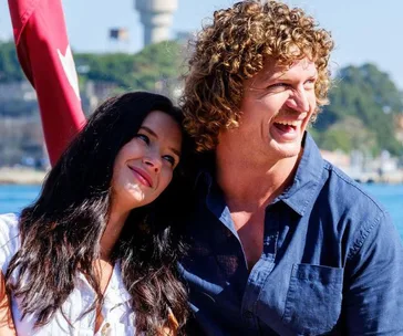 A man and woman smiling together outdoors on a sunny day near the water, with trees in the background.