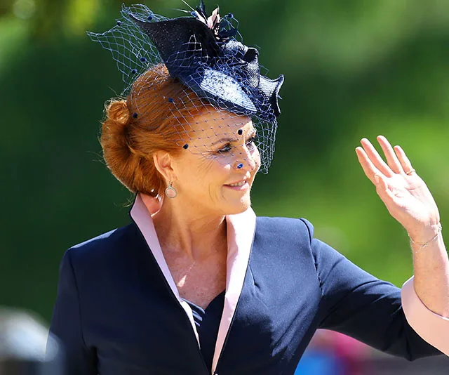 Sarah Ferguson waving in a navy outfit with a matching fascinator and veil, set against a green blurred background.
