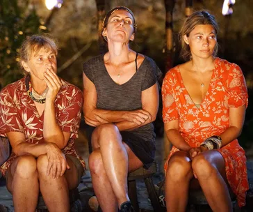 Three women sitting on stools with serious expressions in a dimly lit tribal council setting on Survivor.