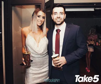 A smiling woman in a silver dress and a man in a suit hold drinks at a party, with "Take 5" in the corner.