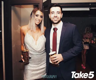 A smiling woman in a silver dress and a man in a suit hold drinks at a party, with "Take 5" in the corner.