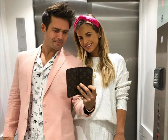 A couple taking a mirror selfie in an elevator, man in a pink blazer, woman in white with a pink headband.