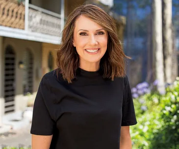Alt text: "Woman smiling outdoors in a black shirt with blurred house and garden background."