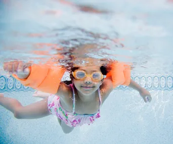 Child swimming underwater in a pool, wearing orange floaties and goggles.