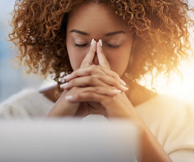 A woman with curly hair sits with eyes closed, fingers pressed together, looking stressed or focused.