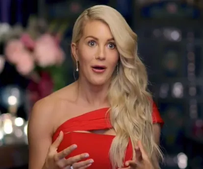 A woman in a red dress with long blonde hair looks surprised, gesturing with hands, blurred background.