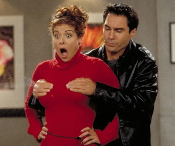 Grace in a red sweater looks shocked as Will, in a black jacket, stands behind her, placing his hands humorously on her chest.