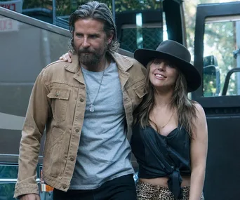 A scene from "A Star is Born" showing a man in a tan jacket and a woman in a black hat, walking closely together.