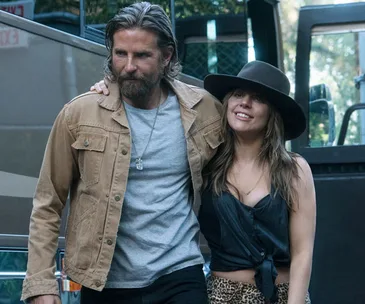 A scene from "A Star is Born" showing a man in a tan jacket and a woman in a black hat, walking closely together.