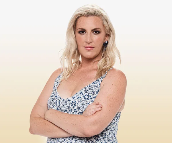 Blonde woman in a patterned dress with arms crossed, against a gradient background.
