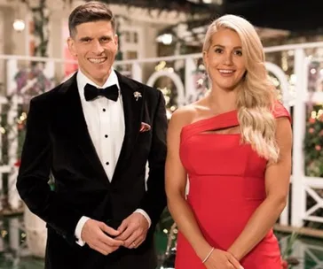 Man in a black tuxedo and woman in a red dress smiling, standing in front of a festive backdrop.