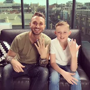 Two people on a couch showing their painted nails for the Polished Man campaign, with a city view behind them.