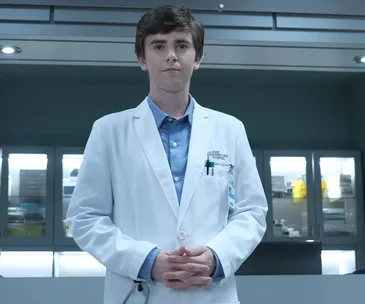 Doctor in a white coat stands in a hospital setting with hands clasped.