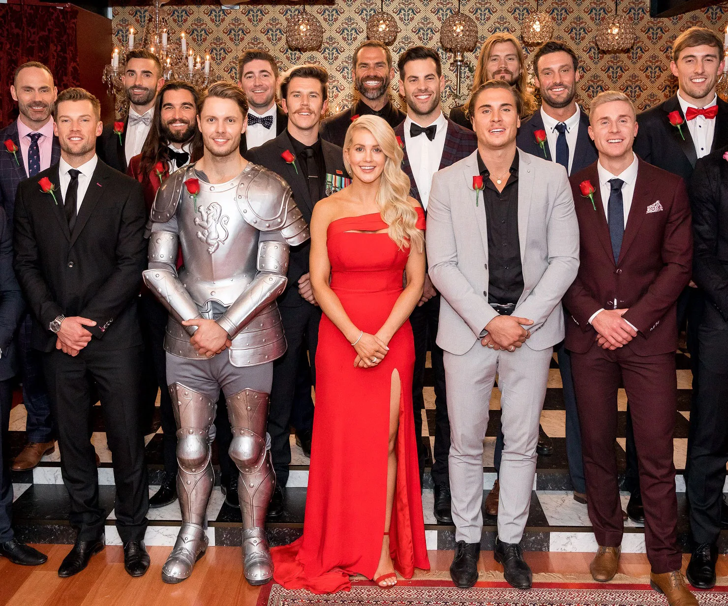 Group photo of "The Bachelorette Australia 2018" contestants in formal wear, with one in a knight costume, posing indoors.